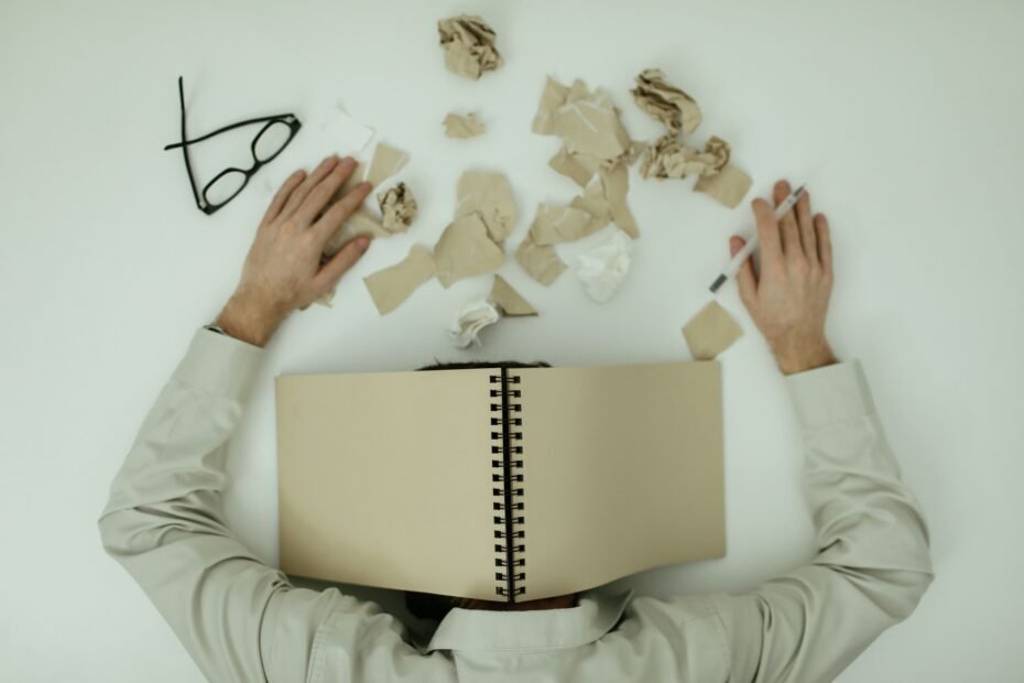 A man is lying on the floor with a notebook over his head and several wadded up papers scattered about on the floor. His glasses also lay on the floor.